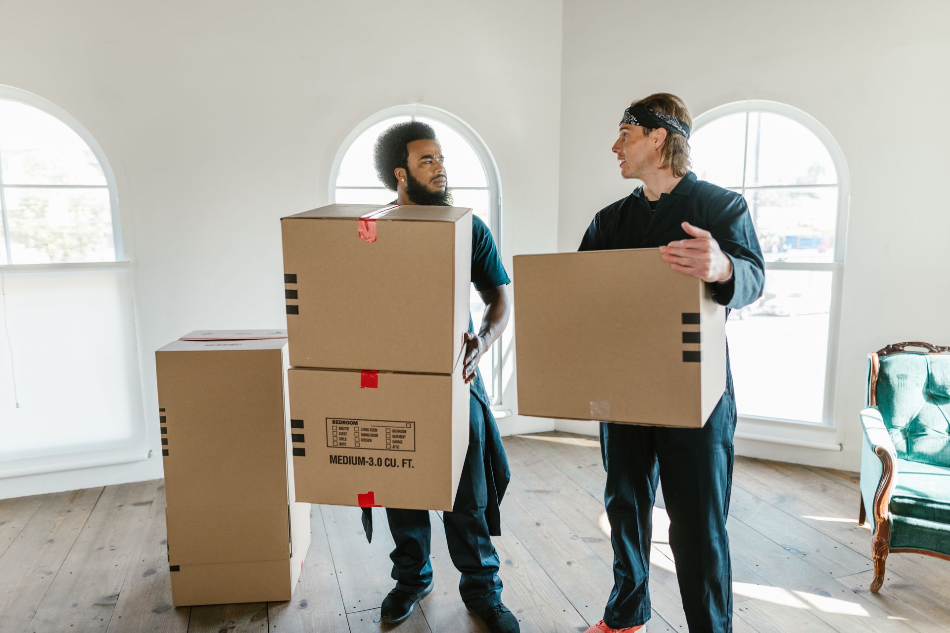 Two professional movers carrying boxes through a bright room with arched windows